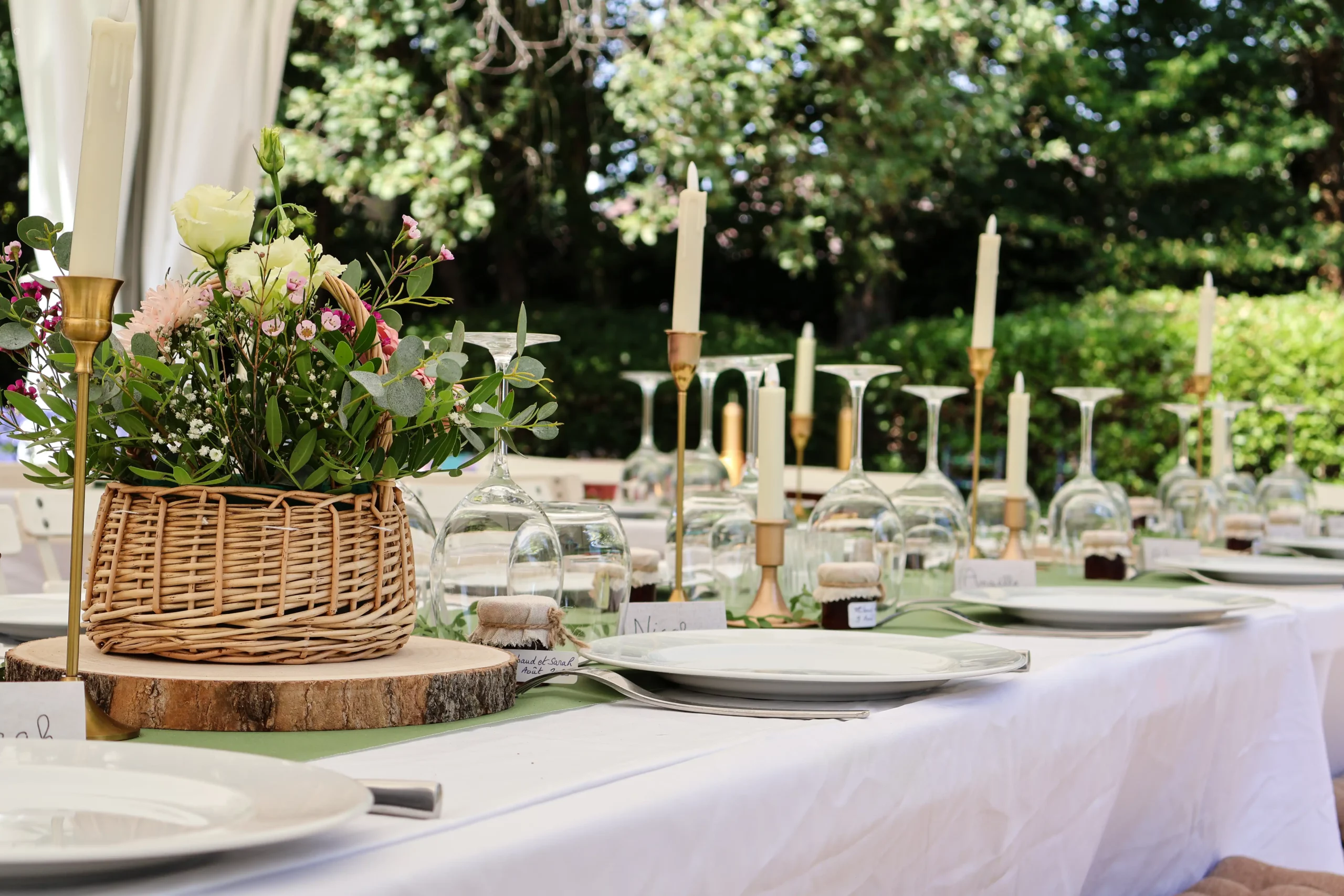 Table de réception dressée en extérieur, panier en osier fleuri, bougies sous cloches en verre, vaisselle blanche sur nappe claire
