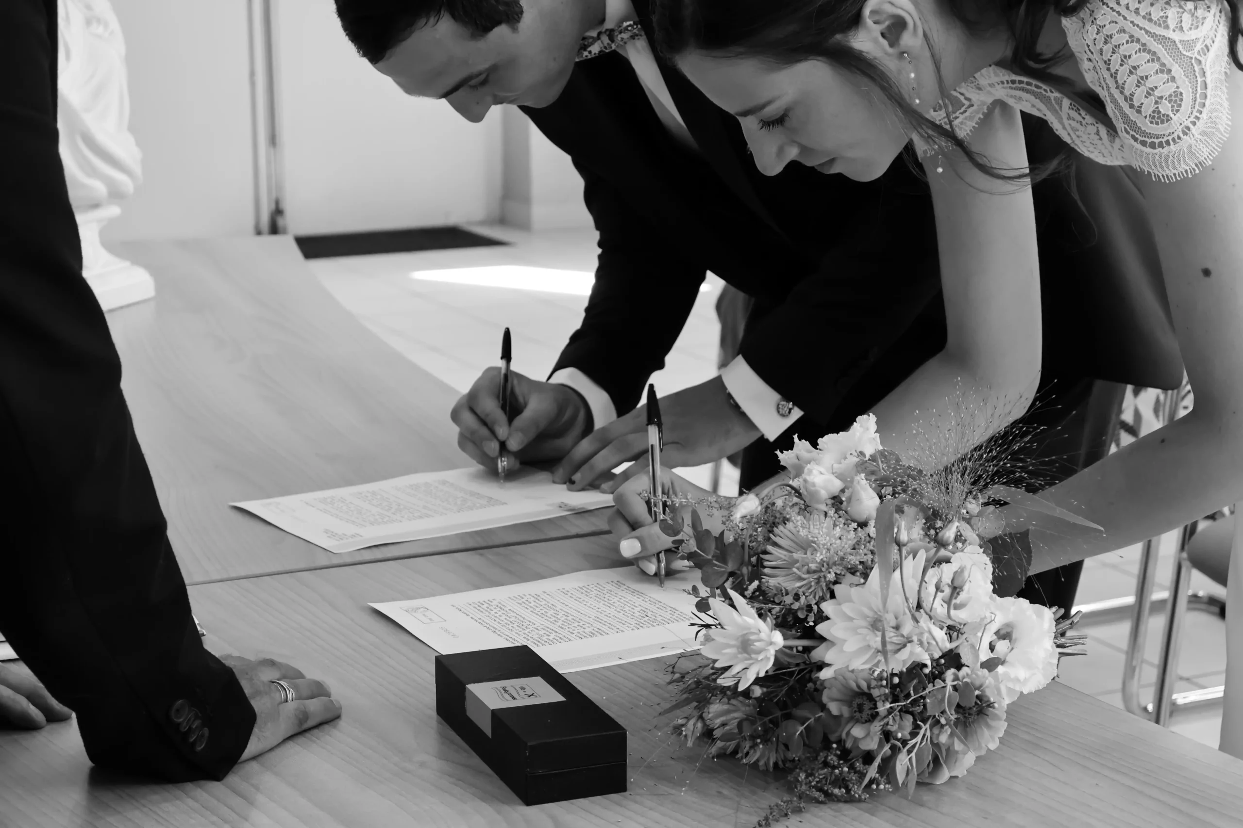Gros plan noir et blanc sur les mains des mariés signant le registre, bouquet posé sur la table
