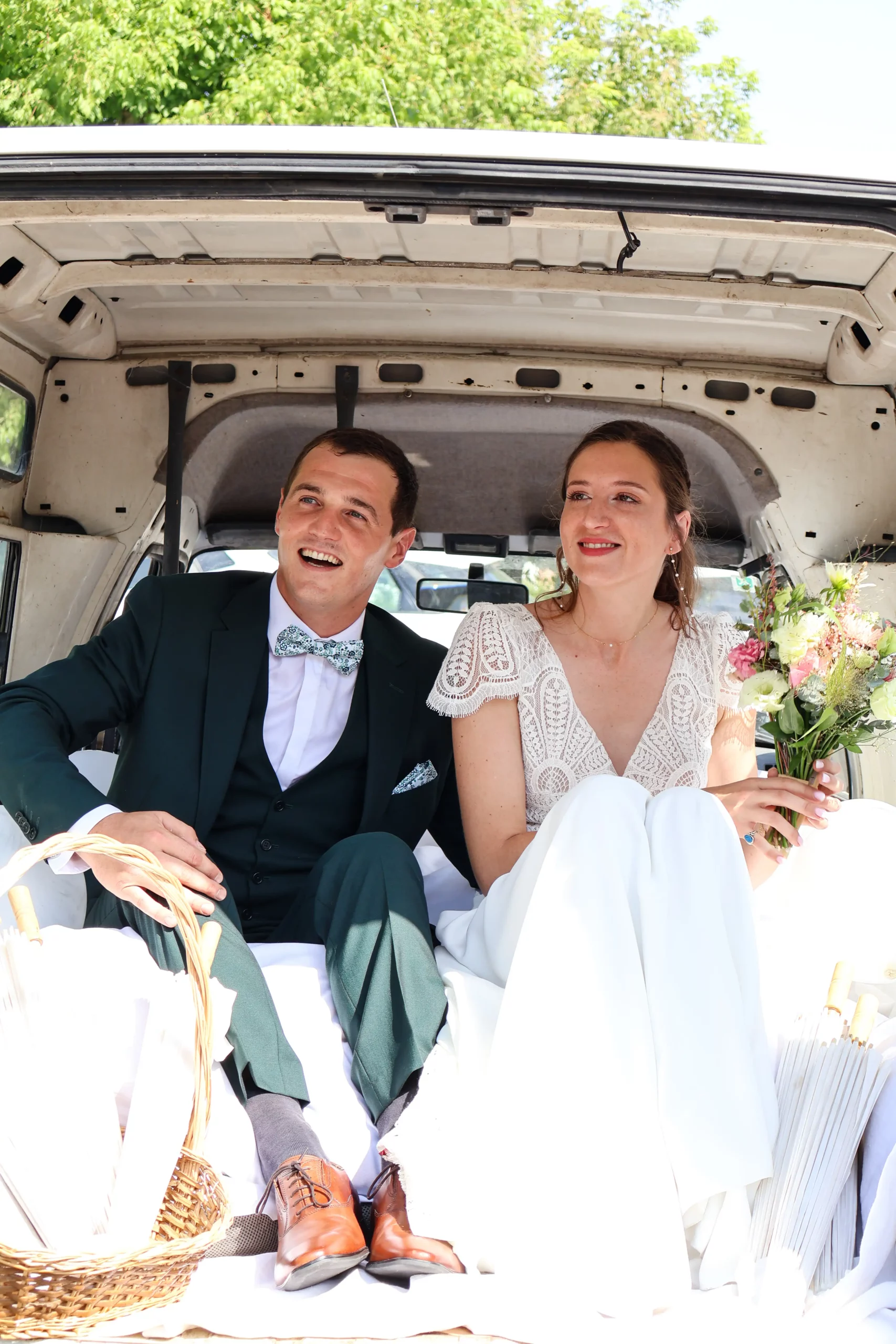 Mariés souriants assis à l'arrière d'un fourgon vintage, lui en costume vert sapin, elle en robe de dentelle avec un bouquet de fleurs